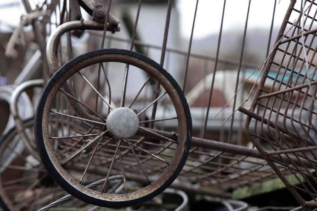 schrotthändler hannover Nahaufnahme eines rostigen, alten Einkaufswagenrads und -rahmens, mit Teilen anderer Einkaufswagen im Hintergrund. Die Speichen des Rads sind intakt, aber die Metalloberflächen weisen Abnutzungs- und Rostspuren auf. Es ist, als wären sie auf einem Schrottplatz in der Nähe vergessen worden. Der unscharfe Hintergrund lenkt den Fokus auf das Rad und den Einkaufswagenrahmen. metallschrott hannover