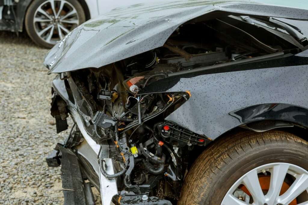 schrotthändler hannover Ein schwarzes Auto mit erheblichen Schäden an der Front, darunter eine zerdrückte Motorhaube und freiliegende Innenteile nach einem Aufprall. Das Fahrzeug, das wahrscheinlich für Schrottpreise Hannover bestimmt ist, steht auf einer Schotterfläche. Im Hintergrund ist teilweise ein anderes Auto zu sehen. metallschrott hannover