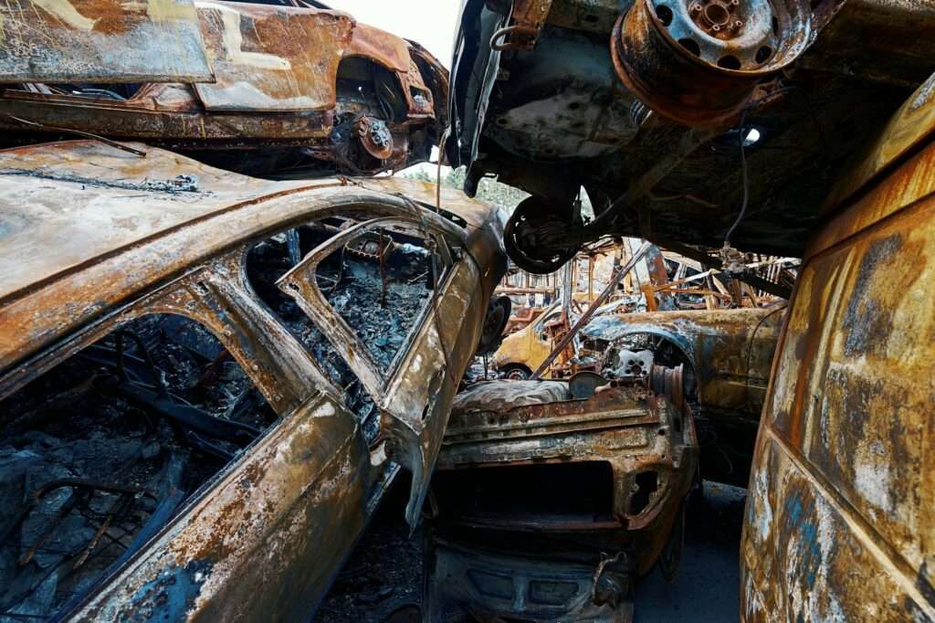 schrotthändler hannover Ein Haufen stark verrosteter, ausgebrannter Autos ist in chaotischer Anordnung übereinander gestapelt. Die Fahrzeuge sind schwer beschädigt, mit sichtbaren Verkohlungen und fehlenden Teilen, was auf einen erheblichen Brandvorfall schließen lässt. Im Hintergrund sind weitere Wrackteile zu sehen, die wahrscheinlich von einem nahegelegenen „Schrotthändler in der Nähe“ gehandhabt wurden. metallschrott hannover