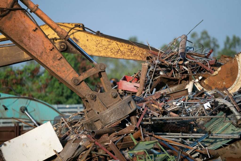 schrotthändler hannover Man sieht einen Baggerarm, der sich durch einen großen Haufen gemischten Schrotts und Schutts arbeitet, darunter verschiedene Stangen, Bleche und Baumaterialstücke. Vor dem Hintergrund von Bäumen und einem klaren Himmel unterstreicht diese Szene die Bedeutung der Schrottpreise Hannover bei der Bewertung von Schrottmaterialien. metallschrott hannover