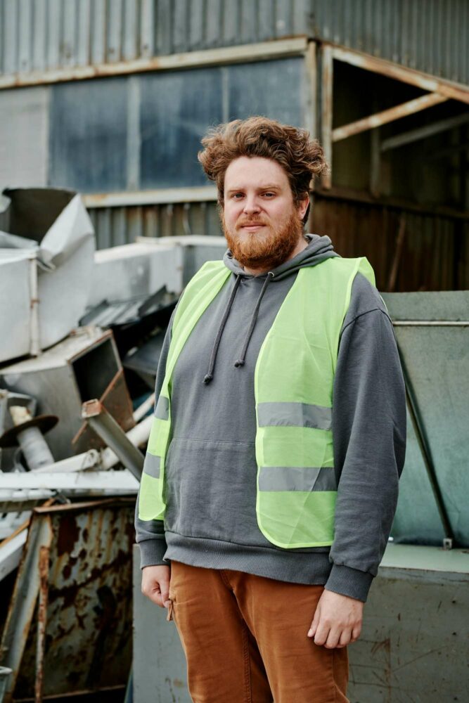 schrotthändler hannover Ein Mann mit Bart steht vor einer Industrieanlage. Er trägt einen grauen Kapuzenpullover und eine neongrüne Warnweste. Im Hintergrund sind Berge von Metallschutt und ein Wellblechgebäude zu sehen. Er hat einen neutralen Gesichtsausdruck und blickt in die Kamera. metallschrott hannover