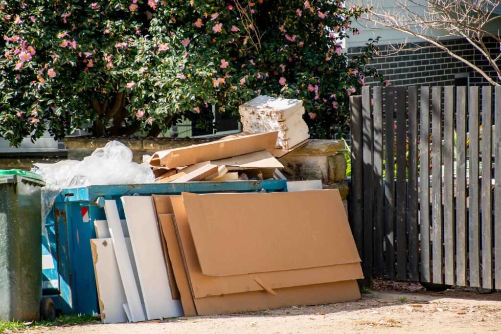 schrotthändler hannover Ein Stapel weggeworfener Kartons und Verpackungsmaterialien, möglicherweise aus einer Haushaltsauflösung Hannovers, ist neben einer blauen Recyclingtonne auf dem Bürgersteig gestapelt. Eine grüne Mülltonne steht in der Nähe, eingerahmt von einem Holzzaun und einem blühenden Baum mit rosa Blüten im Hintergrund. metallschrott hannover