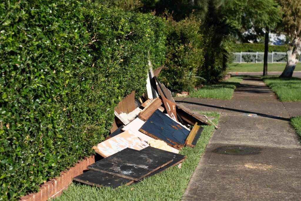 schrotthändler hannover Auf einem Grünstreifen neben dem Bürgersteig, gesäumt von einer üppigen Hecke, wartet ein Haufen ausrangierter Holzbretter und Schutt auf Sperrmüll Abholung Hannover. Die Szene spielt im Freien, im Hintergrund sind Bäume und ein Zaun zu sehen, bereit für die ordnungsgemäße Sperrmüllentsorgung. metallschrott hannover